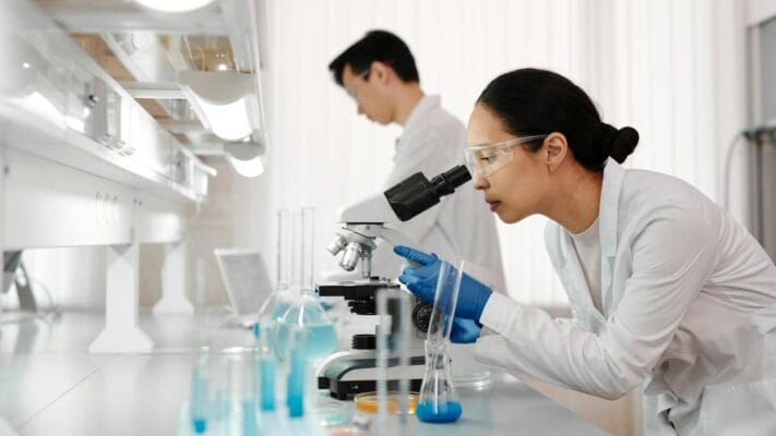Research-grade peptide reconstitution scene — scientist with microscope and blue glassware vials in a clean modern lab
